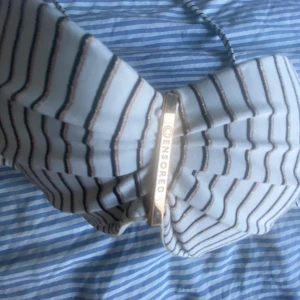 Randig bikiniöverdel från Censored - Supersnygg bikiniöverdel från Censored i vitt med tunna svarta ränder. Bikinin har vadderade kupor och knyt i nacken för perfekt passform. Metallogo framtill ger en extra touch. Perfekt för stranden eller poolen i sommar! Ny och endast provad( ny utan prislapp), storlek 75B