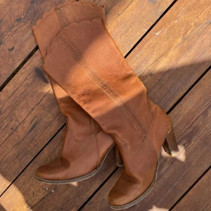 Bruna höga boots med klack - Snygga bruna boots i skinn med hög klack och dekorativa sömmar. Skaftet är högt med vågig kant upptill och diskret mönster i lädret. Perfekta för dig som vill ha en westerninspirerad look med lite extra höjd.