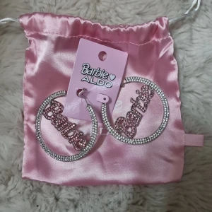 Barbie x ALDO stora creolörhängen - Stora unika creolörhängen från Barbie x ALDO med glittrande stenar i silver och rosa. Ordet 'Barbie' i rosa stenar pryder mitten av örhängena. Kommer med en lyxig rosa satinpåse. Helt nya I förpackning,  köpta från Zalando.