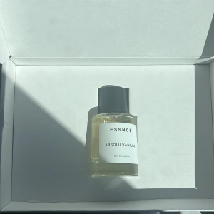 ESSNCE Absolu Vanilla parfym - Minimalistisk parfymflaska från ESSNCE med doften Absolu Vanilla. Flaskan är genomskinlig med ljusgul vätska och har en stilren vit etikett samt grått lock. Perfekt för dig som gillar söta och mjuka vaniljtoner.
