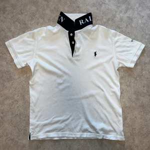 Vit Polo Ralph Lauren t-shirt med tryck - Vit t-shirt från Polo Ralph Lauren med korta ärmar och klassisk pikékrage i svart med stor vit logotyptext. Broderad logga på bröstet och tryckt Ralph Lauren-text på ärmen. Tillverkad i mjuk bomull, perfekt för en clean och sportig look.