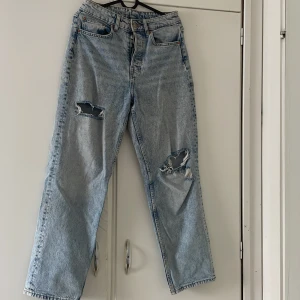 Ljusblå ripped jeans från Divided - Säljer ett par ljusblå jeans från Divided i storlek 36. De har raka ben, hög midja och slitna detaljer med stora hål på båda benen. Klassisk femficksmodell med knappgylf. Perfekta för en avslappnad och trendig look.