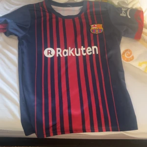 FC Barcelona randig matchtröja - Säljer en officiell FC Barcelona fotbollströja med Rakuten och UNICEF-tryck. Tröjan är blå och röd med smala vertikala ränder, klubbmärke på bröstet och korta ärmar. Tillverkad i lätt och ventilerande material, perfekt för träning eller match.