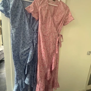 2 st Blommiga omlottklänningar Vero Moda XS - Två långklänningar från Vero Moda i omlottmodell med volangkant och knyt i midjan. En är blå med blommigt mönster, den andra rosa med blommigt mönster. Båda har kort ärm och v-ringning, perfekta för sommarens alla tillfällen.