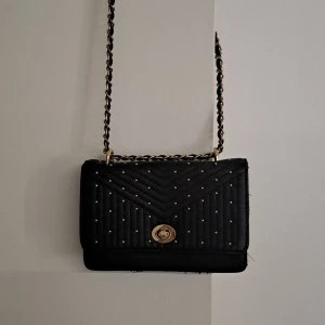 Svart quiltad axelväska med guldkedja - Snygg svart axelväska med quiltad framsida och små guldfärgade nitar. Väskan har en kedjerem i guld och svart som går att justera storleken på, samt ett vridlås i guld. Insidan har kortfack och dragkedja. Perfekt för att lyfta din outfit.