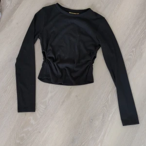 Svart långärmad croppad topp - Svart långärmad topp med croppad passform och håll i sidorna som skapar en snygg effekt. Toppen har rund halsringning och är tillverkad i ett mjukt, stretchigt material som känns bekvämt mot huden. Perfekt för att lyfta din outfit med en enkel men trendig vibe.