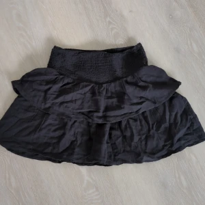 Svart volangkjol från Gina Tricot - Supersöt svart kortkjol från Gina Tricot med bred smock i midjan och dubbla volanger. Kjolen är luftig och har en flowy känsla, perfekt till sneakers eller boots. Enkel att matcha med crop top eller hoodie.