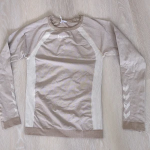 Beige långärmad träningströja Hummel - Sportig beige långärmad tröja från Hummel med vita detaljer längs sidorna och ärmarna. Tröjan har klassiska Hummel-pilar på ärmarna och är tillverkad i en mjuk mix av polyester, polyamid och elastan. Perfekt för träning eller chill dagar.