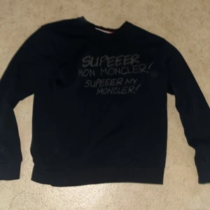 Svart tröja från Moncler - Svart sweatshirt från Moncler med tryckt text på bröstet och klassisk logga broderad på ärmen. Tröjan har rund halsringning och långa ärmar. Tillverkad i mjuk bomull för en skön och avslappnad stil.