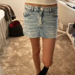 Ljusblå jeanskjol med fickor - Säljer en ljusblå jeanskjol i kort modell med klassisk femficksdesign och bälteshällor. Kjolen har en snygg tvättad look och stängs med knapp och dragkedja framtill. Perfekt för dig som gillar en avslappnad och trendig stil.