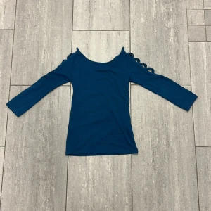 Blå topp med snörning från Amisu - Säljer en snygg blå topp från Amisu i storlek S. Toppen har långa ärmar och coola snörningsdetaljer på axlarna som ger en unik look. Materialet är mjukt och stretchigt, perfekt för dig som gillar stilrena men ändå speciella plagg.