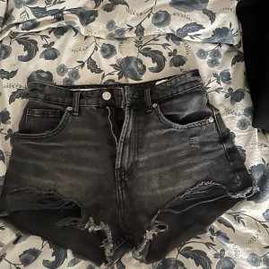 Svarta jeansshorts från Zara - Snygga svarta jeansshorts från Zara med hög midja och råa, fransiga kanter. Klassisk femficksmodell med dragkedja och knapp framtill. Perfekta för en cool och avslappnad stil under sommaren.