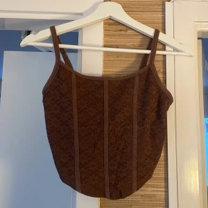 Brun korsettopp från Hollister S - Supersnygg brun korsettopp från Hollister med smala axelband och broderade detaljer framtill. Toppen har en croppad passform och är tillverkad i bomullsmix med justerbara axelband. Perfekt att styla med jeans eller kjol för en trendig look.