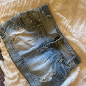 Ljusblå jeanskjol med slitningar - Snygg ljusblå jeanskjol med slitna detaljer och råa kanter. Klassisk femficksmodell, bälteshällor och dubbla knappar framtill.