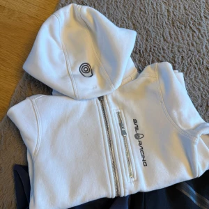 Vit hoodie från Sail Racing med dragkedja - Vit hoodie från Sail Racing med broderad logga på huvan och bröstet. Tröjan har en snygg half zip-dragkedja och en extra dragkedjeficka framtill. Mjuk bomullskänsla och huva för chill stil.