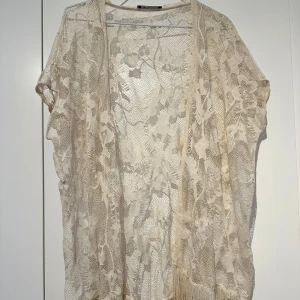 Cardiga/Kimono - Säljer en superfin beige overshirt i spets från Ginatricot. Den har blommigt mönster, korta ärmar och långa fransar längst ner. Perfekt att slänga över en topp för en boho-vibe. Lätt och luftig, passar grymt till sommaren!