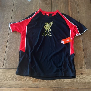 Liverpool FC tröja 2019 fanwear - Liverpool FC fotbollströja i svart med röda ärmar och vita detaljer. Stort L.F.C.-tryck i guld på bröstet. Tillverkad i lätt och ventilerande polyester, perfekt för träning eller match. Klassisk passform med korta ärmar och rund hals.  Helt nytt skick och lappen och tagen sitter även kvar!