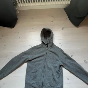 Grå hoodie från Polo Ralph Lauren - Klassisk grå hoodie från Polo Ralph Lauren med dragkedja framtill, huva och två fickor. Tröjan har det ikoniska broderade logotypen på bröstet och är tillverkad i mjuk bomullsmix. Andvänd rätt många ggr med fortfarande väldigt fint skick!