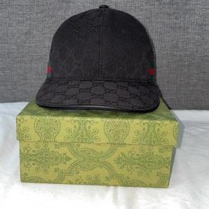 Svart keps från Gucci med GG-mönster - Snygg svart keps från Gucci med diskret GG-monogram över hela kepsen. Klassisk böjd skärm och justerbart spänne bak i svart läder. Sidorna har Guccis ikoniska gröna och röda ränder. Tillverkad i bomull och detaljer i läder.