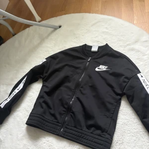 Svart mjukisett från Nike - Svarta mjukisbyxor från Nike med elastisk midja och smal passform. Byxorna har ribbade muddar vid bensluten och är tillverkade i ett mjukt och bekvämt material. Perfekta för chill eller träning, med klassisk Nike-stil. Kommer ihop med en svart sip hoodie från Nike, kommer som ett sett