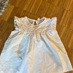 Vit tröja  med spetsdetaljer - Söt blus med broderad spets upptill och volangärmar. Tröjan har en luftig passform och är ärmlös, perfekt för varma dagar. Knäppning bak i nacken och lätt A-linjeformad siluett. Tillverkad i bomull för en mjuk och bekväm känsla.