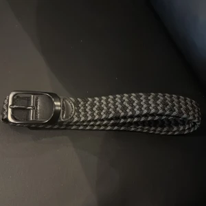 Svart och grått flätat bälte - Snyggt flätat bälte i svart och grått med klassiskt spänne i metall och detaljer i skinn. Bältet har ett coolt zigzag-mönster och passar perfekt till jeans eller chinos för en stilren look.