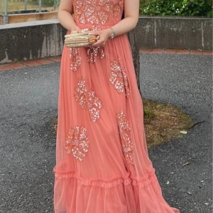 Lång korallrosa balklänning med paljetter - Superfin lång klänning i korallrosa med glittriga paljettmönster. Klänningen har ärmlös design, markerad midja och volangdetaljer längst ner. Perfekt för bal eller fest, ger en riktigt glam känsla med sitt flowiga tyg och skimrande detaljer. Passar xs och s. 