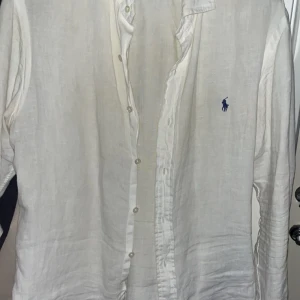 Vit linneskjorta från Ralph Lauren - Vit långärmad skjorta i linne från Ralph Lauren med klassisk krage och knäppning framtill. Skjortan har den ikoniska blå broderade loggan på bröstet och en avslappnad passform. Perfekt för en clean och stilren look.