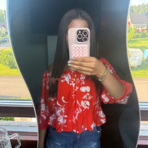 Röd blommig offshoulder blus - Superfin röd blus med vita blommor och offshoulder-modell. Blusen har trekvartsärmar, knappar framtill och smockad midja som ger en snygg volangkant. Perfekt för sommarens alla tillfällen och riktigt skön att bära.