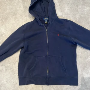 Marinblå hoodie från Polo Ralph Lauren - Marinblå hoodie från Polo Ralph Lauren med dragkedja och huva. Klassisk röd broderad logga på bröstet och stora fickor framtill. Perfekt för en chill och stilren look. Tillverkad i mjuk bomull som känns skön mot huden.