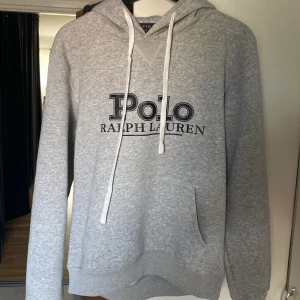 Grå hoodie från Polo Ralph Lauren - Säljer en grå hoodie från Polo Ralph Lauren med stor logga framtill. Tröjan har huva med vita snören, känguruficka och ribbade muddar. Perfekt för dig som gillar en clean och klassisk streetstil.