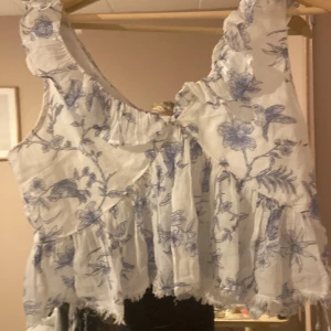 Vit blommig volangtopp med blått mönster - Supersöt vit topp med blått blommönster och volangdetaljer. Ärmlös modell med rynkade kanter och lätt ruffig nederkant. Perfekt för en somrig look och riktigt skön att bära. Luftigt material och romantisk vibe.