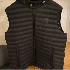 Svart dunväst Moose Knuckles - Säljer en svart dunväst från Moose Knuckles med horisontella quiltade sömmar och huva. Västen har två dragkedjeförsedda fickor framtill och en diskret logotyp på bröstet. Perfekt för lager-på-lager och snygg till streetwear.