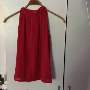 Röd blus - Supersöt röd blus i luftig polyester med ärmlös design och snygg knytning i nacken. Blusen har en lös passform och faller fint, perfekt för dig som vill sticka ut med en poppig färg och enkel stil.