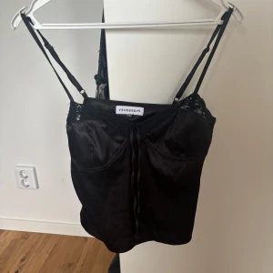 Svart satinlinnetopp från Venderby's - Supersnygg svart linnetopp från Venderby's i glansigt satinmaterial med tunna axelband, spetsdetaljer upptill och en liten knytning framtill. Perfekt för att lyfta vilken outfit som helst med en chic vibe.