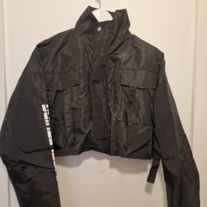Cool svart vindjacka  - Svart croppad vindjacka från PrettyLittleThing med hög krage, dragkedja. Jackan har flera stora fickor framtill och coola detaljer. Perfekt för dig som gillar streetstyle och vill sticka ut.
