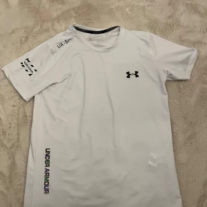 Vit Under Armour t-shirt i polyester - Snygg vit t-shirt från Under Armour med svart logga på bröstet och tryck på ärmen samt längs sidan. Tillverkad i mjuk och lätt polyester, perfekt för träning. Klassisk passform med korta ärmar och rund halsringning. Den är reflektiv