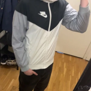 Nike vindjacka i svart och grått - Nike vindjacka med svart överdel och ljusgrå ärmar. Jackan har dragkedja framtill, vit Nike-logga på bröstet och en sportig look. Perfekt för dig som gillar streetwear och vill ha en lätt jacka med snygg färgblockdesign.