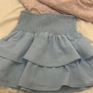 Ljusblå volangkjol från Gina Tricot - Supersöt ljusblå kortkjol från Gina Tricot med bred smock i midjan och dubbla volanger. Kjolen är luftig och har en flowy känsla, perfekt för dig som gillar en feminin och trendig stil.