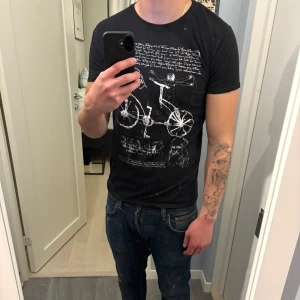 Svart t-shirt med Da Vinci-tryck - Svart t-shirt med coolt vitt tryck inspirerat av Leonardo da Vinci, med skisser, text och cykelmotiv. Klassisk passform och rund hals.