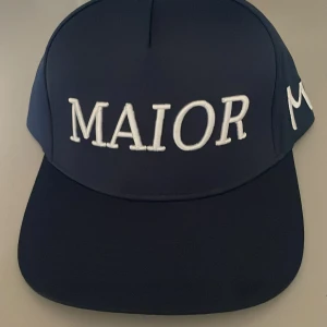 Mörkblå keps MAIOR brodyr - Snygg mörkblå keps med vit brodyrtext 'MAIOR' framtill och siffrorna 20 och 23 broderade på sidorna. Klassisk snapback-modell med böjd skärm och justerbar baksida. Perfekt för dig som vill sticka ut med stilren streetwear.