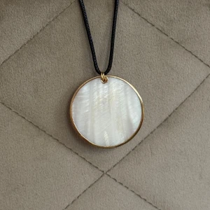 Runt halsband med pärlemor och guldkant - Stilrent halsband med ett runt hänge i snäcka/pärlemor infattad i tunn guldfärgad kant. Hänger i ett svart snöre för en enkel men elegant look, justerbart. Funkar för alla stilar och tillfällen, lätt att matcha med andra smycken 🥰 perfekt för sommaren och stranden! Köp fler smycken och få rabatt på bundle :)