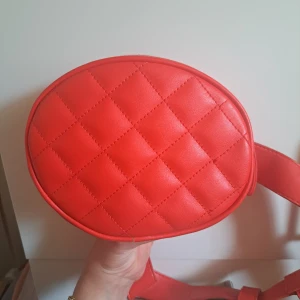 Röd quiltad magväska från Monki - Säljer en rund, quiltad magväska från Monki i stark röd färg. Väskan har bredt bälte och dragkedja upptill. Tillverkad i syntetmaterial och perfekt för att bära det viktigaste. Enkel och snygg accessoar som sticker ut.