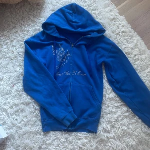 Blå hoodie med strassängel och text - Blå hoodie med dragkedja och huva. På framsidan finns en strassdekorerad ängel med pilbåge och texten 'Best Yet To Come' i glittriga stenar. Mjuk bomullskänsla och långa ärmar. Perfekt för dig som gillar statement-plagg med lite extra bling.