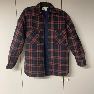 Rutig overshirt från JoeY, marinröd - Snygg overshirt från JoeY i röd och mörkblå rutig design. Skjortan har två bröstfickor med knappar, klassisk krage och långa ärmar. Perfekt att bära öppen över en t-shirt eller stängd som skjorta. Materialet är mjukt och skönt, troligtvis bomull.