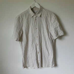 Randig kortärmad skjorta från Zara - Snygg kortärmad skjorta från Zara i beige med tunna svarta och blå ränder. Skjortan har klassisk krage, knäppning framtill och är tillverkad i en mix av bomull, polyester, linne och viskos. Perfekt för en chill och stilren look. Artnr: C378