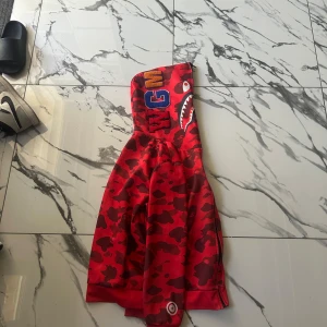 Röd camo bomberjacka från BAPE - Säljer en ikonisk röd camo bomberjacka från BAPE med hajmotiv på huvan och broderade bokstäver. Jackan har dragkedja och coola detaljer som gör den till ett statement-plagg. Perfekt för dig som vill sticka ut med streetwear-stil.