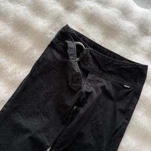 Svarta bootcut-byxor från Bench - Svarta bootcut-byxor från Bench med bred linning och dekorativt bälte med metallspänne. Byxorna har en snygg utsvängd passform och Bench-logga baktill i midjan. Perfekta för dig som gillar en avslappnad men trendig stil.