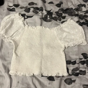 Vit blus med puffärm och broderi - Supersöt vit blus med smockad kropp och puffiga korta ärmar i broderad bomull. Ärmarna har volangkant och blommiga hålbroderier som ger en romantisk vibe. Toppen är croppad och har volangdetaljer längs halsringning och nederkant.