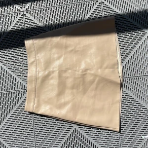 Beige skinnkjol i kort modell - Snygg beige kortkjol i skinnimitation med slät yta och enkel design. Kjolen har en rak passform och är fodrad på insidan för extra komfort. Perfekt att styla med boots eller sneakers för en trendig look.